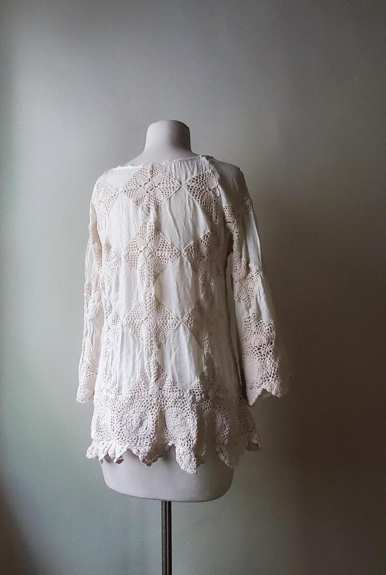 May include: A white, long-sleeved top with intricate crochet detailing. The top features a round neckline, bell sleeves, and a scalloped hem. The crochet design is a lighter shade of white, creating a subtle contrast. The garment is displayed on a mannequin.