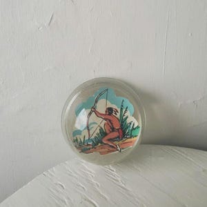 Può includere: Una cupola di plastica trasparente con un'immagine colorata di un indigeno con arco e freccia in un paesaggio verde e blu.