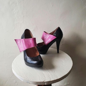 May include: A pair of black high-heeled Mary Jane shoes with a pink strap. The shoes have a platform sole and a stiletto heel. The shoes are displayed on a white, round table. The heels are approximately 10 cm high.