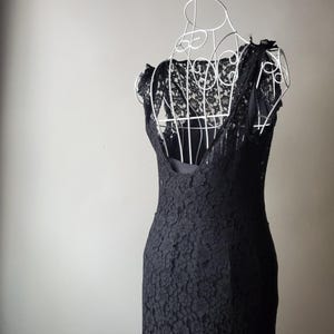 May include: A black lace dress displayed on a white wire dress form. The dress features a floral lace pattern and a scoop neckline. The dress is sleeveless and has a fitted silhouette, ideal for formal occasions.