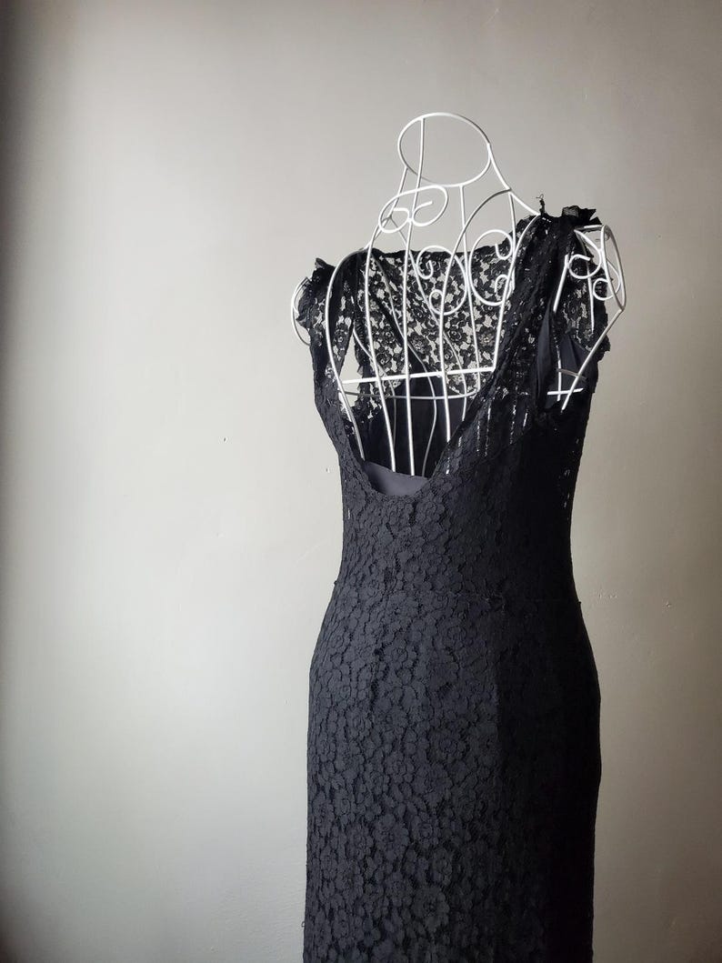 May include: A black lace dress displayed on a white wire dress form. The dress features a floral lace pattern and a scoop neckline. The dress is sleeveless and has a fitted silhouette, ideal for formal occasions.