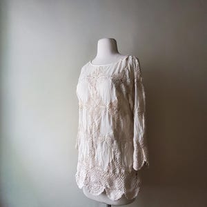 May include: Off-white, long-sleeved blouse with crochet detailing. The blouse features a round neckline and a scalloped hem. The crochet work is visible on the sleeves and the lower portion of the blouse. The blouse is displayed on a mannequin.