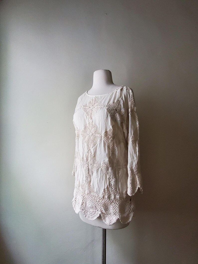 May include: Off-white, long-sleeved blouse with crochet detailing. The blouse features a round neckline and a scalloped hem. The crochet work is visible on the sleeves and the lower portion of the blouse. The blouse is displayed on a mannequin.