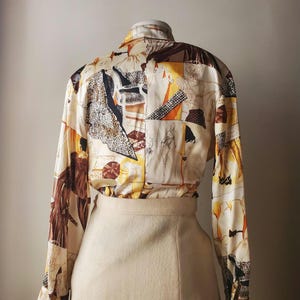 Puede incluir: Una blusa vintage de manga larga con un dise&ntilde;o de patchwork &uacute;nico. La blusa presenta un fondo color crema con varias ilustraciones abstractas y de figuras en tonos marr&oacute;n, negro, naranja y amarillo. Se combina con una falda beige.