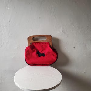 May include: A red handbag with a wooden handle and a black Scottie dog silhouette. The bag has a rounded shape and a black trim. The handle is made of light-colored wood. The bag is sitting on a white, round table.