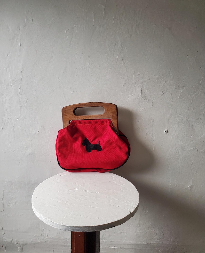 May include: A red handbag with a wooden handle and a black Scottie dog silhouette. The bag has a rounded shape and a black trim. The handle is made of light-colored wood. The bag is sitting on a white, round table.