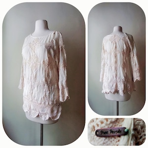 May include: Cream-colored long-sleeve top with intricate crochet detailing. The top features a round neckline, three-quarter sleeves, and a scalloped hem. The design includes floral and geometric patterns. The tag reads "Free People".