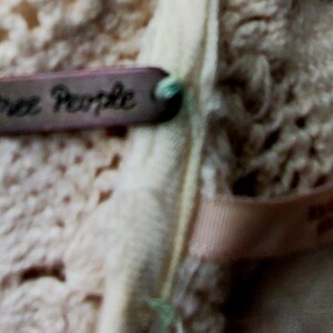 May include: Close-up of a cream-colored crocheted garment with a brown tag that reads "Free People." The tag is attached with a green string. A light pink ribbon with text is also visible.