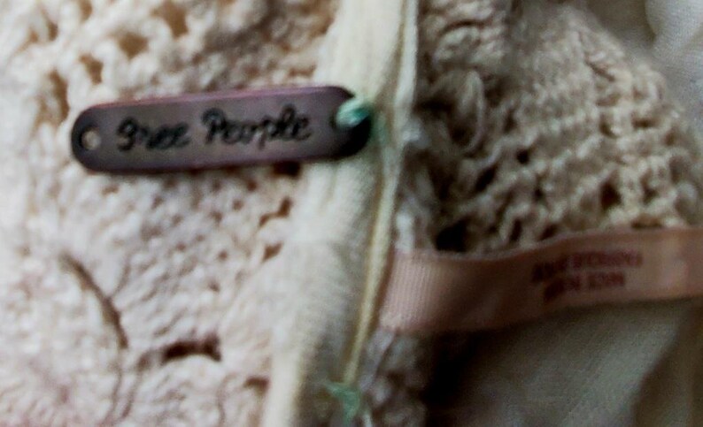 May include: Close-up of a cream-colored crocheted garment with a brown tag that reads "Free People." The tag is attached with a green string. A light pink ribbon with text is also visible.