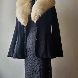 May include: A black velvet jacket with a cream-colored fur collar hangs over a black lace dress. The jacket has a flared silhouette and two buttons. The dress has a floral lace pattern and a ruffled hem. The outfit is displayed on a white hanger.