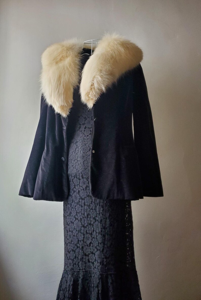 May include: A black velvet jacket with a cream-colored fur collar hangs over a black lace dress. The jacket has a flared silhouette and two buttons. The dress has a floral lace pattern and a ruffled hem. The outfit is displayed on a white hanger.
