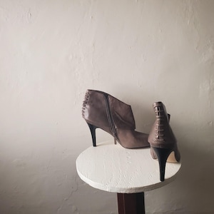 May include: A pair of brown ankle boots with high, black heels. One boot features a side zipper, while the other has a lace-up design on the back. The boots are displayed on a small, white, round table.