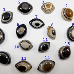 May include: A collection of fifteen oval-shaped agate stones, each with a unique eye-like pattern. The stones feature a variety of colors, including black, white, brown, and cream, with concentric circles and bands. The stones are arranged on a white surface.