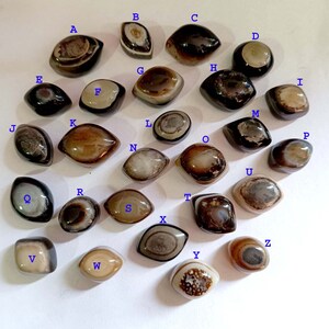 May include: Twenty-six agate stones, each with a unique pattern of brown, black, and white, arranged in alphabetical order on a white surface.