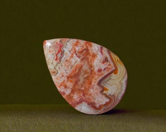 May include: A polished, teardrop-shaped gemstone with a striking pattern of red, orange, and white swirls and bands. The stone has a smooth, glossy surface and is set against a dark green background.
