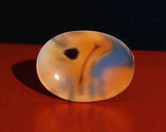 Pode incluir: Pedra preciosa em forma oval com superfície polida. A pedra apresenta uma mistura de tons laranja, castanho e azul, com uma mancha escura proeminente. A pedra preciosa está sobre uma superfície vermelha com fundo preto.