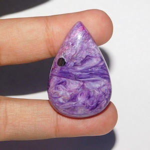 May include: A teardrop-shaped gemstone with a swirling pattern of purple and white. The polished stone has a small, dark hole near the top. The gemstone is held between two fingers against a white background.