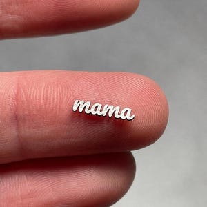 May include: A small, silver-colored charm with the word "mama" in a cursive font. The charm is resting on a fingertip, with a blurred background.