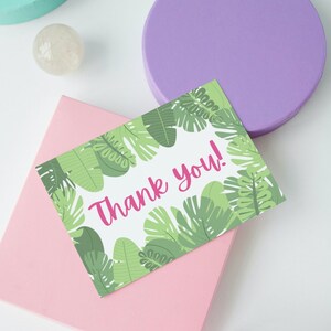 May include: A greeting card with a tropical leaf design and the words "Thank you!" in pink. The card is surrounded by green leaves and sits on a pink envelope. A clear, round object and a purple circle are in the background.