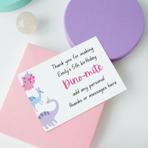 May include: A white card with the text "Thank you for making Emily's 5th birthday Dino-mite" and space for a personal message. The card features cartoon dinosaurs in pink, blue, and purple.