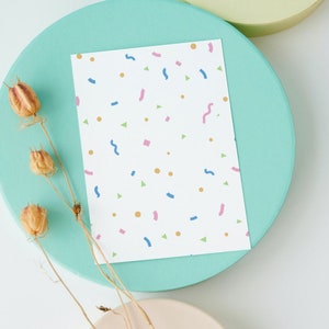 May include: A white card with a confetti pattern in blue, pink, green, and gold. The card is on a light blue circular platform, with dried seed pods and a white background.