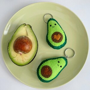 Puede incluir: Un aguacate fresco cortado por la mitad con un hueso marrón grande junto a dos llaveros de fieltro hechos a mano con forma de aguacate. Cada llavero tiene un exterior verde brillante, un borde verde oscuro, un hueso en forma de corazón.