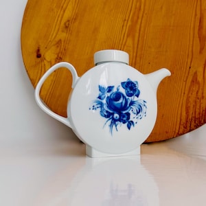Può includere: Teiera in ceramica bianca con un design floreale blu. La teiera ha un corpo rotondo, un manico curvo e un beccuccio. Il design floreale presenta una grande rosa e altri fiori. La teiera è su una superficie bianca con uno sfondo di legno.