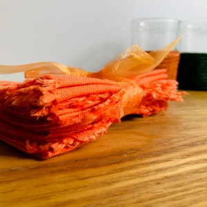 Könnte beinhalten: Ein Stapel orangefarbener Stoffservietten mit ausgefransten Kanten, die mit einem goldenen Band zusammengebunden sind. Die Servietten liegen auf einer Holzoberfläche.