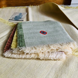 Puede incluir: Un conjunto de posavasos cuadrados con flecos en tonos verdes, rosas y amarillos, con flecos de color claro. Los posavasos están sobre una tela tejida de color amarillo claro. Una etiqueta con el texto "Water Guarantee" es visible.