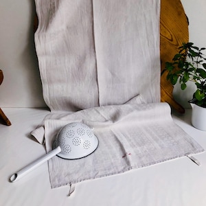 May include: White enamel colander with a long handle, sitting on a white linen tea towel with a red embroidered design. The towel is folded and draped over a wooden surface.