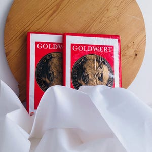 Peut inclure: Deux paquets rouges et blancs avec le mot "GOLDWERT" imprimé. Chaque paquet présente un motif de pièce d'or avec du texte. Les paquets sont sur une surface blanche avec un fond en bois.