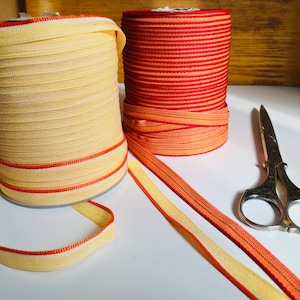 Puede incluir: Dos carretes de cinta, uno amarillo y naranja, el otro rojo y naranja, con un par de tijeras plateadas sobre una superficie blanca.