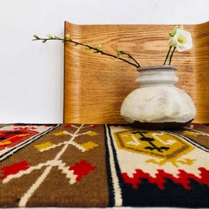 Könnte beinhalten: Eine weiße Keramikvase mit braunem Sockel steht auf einem braunen und weißen gemusterten Teppich. Die Vase ist mit einem Zweig weißer Blumen und grünen Blättern gefüllt. Die Vase steht vor einem Holztablett.