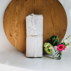 May include: A rolled white fabric bundle tied with twine, set against a large, round wooden backdrop. A small glass jar holds a vibrant floral arrangement with pink, white, and green blooms. The scene is set on a white surface.