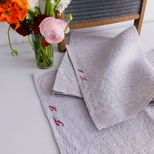 Könnte beinhalten: Drei graue Leinen-Servietten mit roten gestickten Initialen. Die Servietten sind gefaltet und auf einer weißen Oberfläche gestapelt.