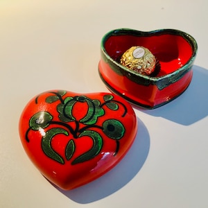 Op de afbeelding: Een rode hartvormige keramische sieradendoos met groene bloemmotieven. De doos is open en bevat een in goud verpakte chocolade.