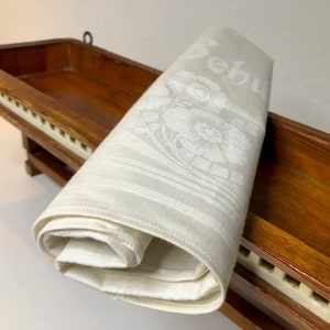 Könnte beinhalten: Ein gefaltetes, cremefarbenes Leinen-Serviette mit floralen und gestreiften Mustern und dem Schriftzug "Bebin". Die Serviette liegt auf einem hölzernen Pflanzkasten mit dunklem Finish und weißem Zierrat.