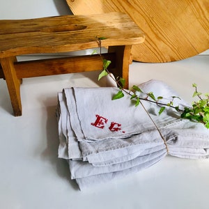 Puede incluir: Un mantel de mesa de lino blanco con bordado rojo que dice "E.G." está doblado y atado con una cuerda. Un pequeño taburete de madera está en el fondo.