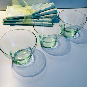 May include: Three clear glass bowls with a light green tint, each with a rounded base and angled sides. A bundle of light green napkins tied with a yellow ribbon is in the background. The bowls cast shadows on the white surface.