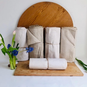 Puede incluir: Varios rollos de tela de lino en varios colores neutros, incluyendo blanco y beige. Los rollos de tela están atados con hilo y dispuestos sobre una tabla de madera. Un jarrón de flores rosas y azules añade un toque de color.