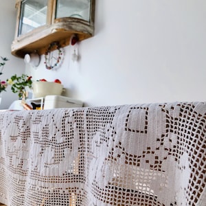 Könnte beinhalten: Weiße Häkeltischdecke mit einem zarten Blumenmuster. Die Tischdecke ist über einen Holztisch gelegt und hängt an den Seiten herunter.