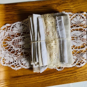 Może przedstawiać: Stosik białych serwetek lnianych z białą koronkową lamówką. Serwetki są złożone i związane razem białą wstążką. Serwetki znajdują się na drewnianej tacy z białą koronkową serwetką.