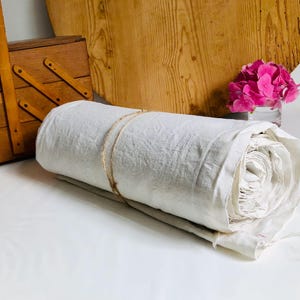 May include: A roll of off-white linen fabric tied with twine. A wooden sewing box and a wooden circular board are in the background. A small glass vase holds pink flowers.