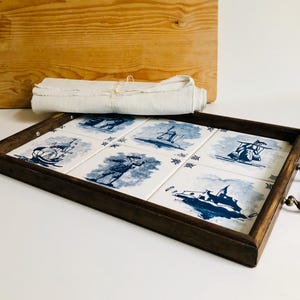 Peut inclure: Un plateau de service rectangulaire en bois avec un cadre marron foncé et des poignées en laiton. Le plateau présente six carreaux blancs avec des illustrations nautiques bleues. Un tissu blanc roulé et une planche à découper en bois sont en arrière-plan.