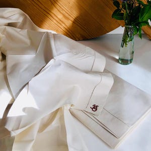 Peut inclure: Ensemble de literie en lin crème avec initiales brodées. L'ensemble comprend un drap et une taie d'oreiller pliés, avec un monogramme rouge foncé. Un petit vase en verre avec de la verdure est à proximité, et une planche à découper en bois est en arrière-plan.