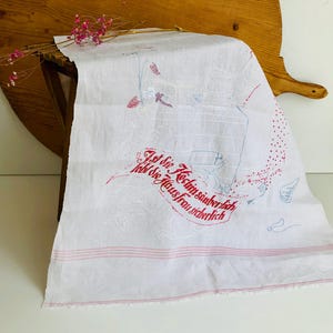 以下が含まれることがあります： 赤と青の刺繍が施された白いリネンのキッチンタオル。赤い文字とキッチン用品のイラストが描かれたバナーが付いています。タオルの下部には2本の赤いストライプがあります。
