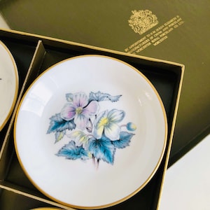 Könnte beinhalten: Set aus weißen Porzellantellern mit einem Blumendesign in Rosa, Gelb und Blau, akzentuiert mit goldenen Rändern. Die Teller sind in einer grünen Box angeordnet, ein klassisches Geschirrset.