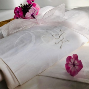 Pode incluir: Um pano de linho branco dobrado com iniciais bordadas, amarrado com uma fita rosa transparente. Uma flor rosa e um cacho de flores rosas são colocados perto do pano, sugerindo um presente ou um artigo decorativo.