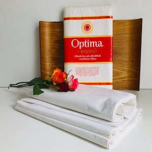 Könnte beinhalten: Ein Stapel weißer Baumwollbettwäsche mit einer roten und weißen Packung "Optima Spezial" Bettwäsche im Hintergrund. Die Packung lautet "2 Bettücher mit allmählich verstärkter Mitte".