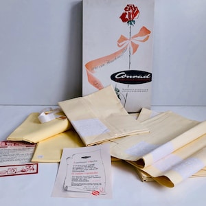 May include: A vintage set of pale yellow linens, including folded sheets and pillowcases, with white lace detailing. The set is packaged with a box featuring a rose illustration and the brand name "Conrad".